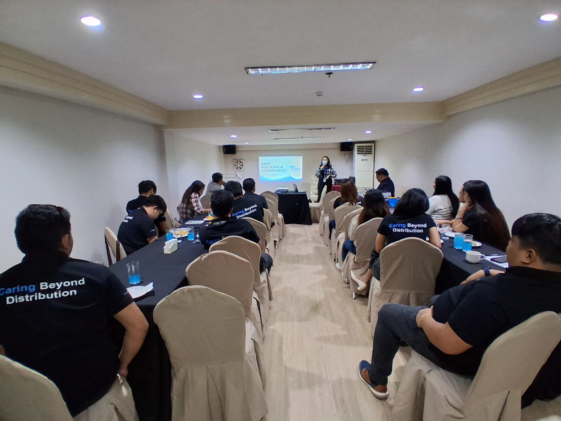 AMZ employees seated around conference tables in "Caring Beyond Distribution" shirts, listening to a speaker presenting the AMZ Policies and Standards slide.