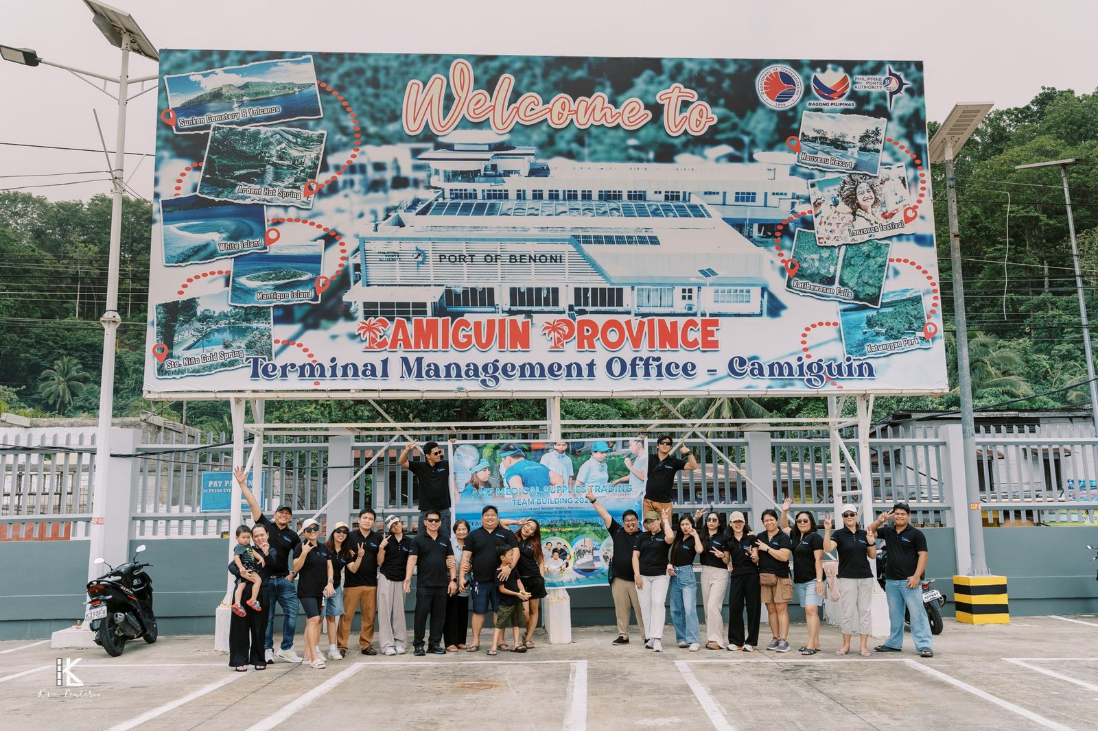 The AMZ team in black shirts holding the event banner beneath the Welcome to Camiguin Province sign at the port