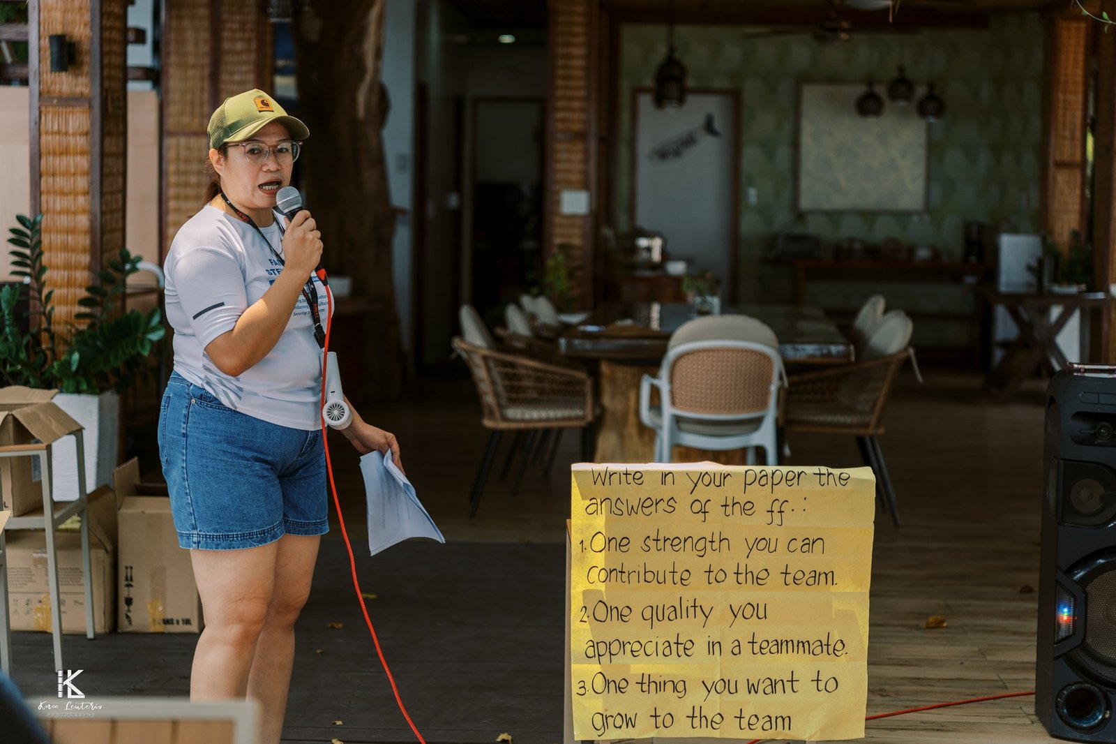 A facilitator speaking into a microphone beside a handwritten sign listing three reflection questions for the team